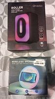Two wireless speakers in original factory packaging on a gray patterned surface. Oraolo Roller speaker box is visible along with a round wireless speaker box featuring an RGB light ring.