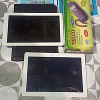 Two white tablets, one with cracked screen showing Yitaogra boot screen and another showing Android boot screen, black case beneath tablets, boxed TELCO telephone handset and two toy tablets