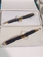 Two black video recording pens with gold accents, one still sealed in plastic inside its box and the other placed in molded tray in its box.
