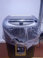 Front and top view of the silver and black paper shredder with plastic waste bin covered by a transparent plastic bag.