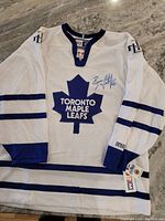 Full view of the white Toronto Maple Leafs jersey signed by Brian Leetch with blue stripes on sleeves and bottom hem.