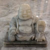 Front view of the white marble Happy Buddha statue showing the Buddha's smiling face, raised belly, and a beaded item in the right hand.