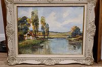 Oil painting of a landscape featuring a small house by a river with tall trees and hills, in an ornate cream-colored frame.