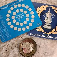Top-down photo of 1970 World Cup coin collection album holder with 30 coins arranged in circular pattern and labeled country names, alongside a blue FA Cup Centennial album holder featuring silver cup image and gold laurel design, with a dish holding loose coins of mixed sizes.
