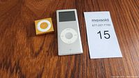 Yellow/Gold and Silver/White Apple iPod Nano devices placed on wooden surface with MaxSold lot card beside them