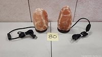 Two Himalayan salt lamps side by side, both about 8 inches tall, with cords and plugs attached.
