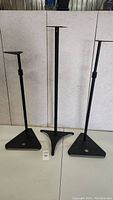 Three black speaker stands, two adjustable, one fixed height, positioned against white background
