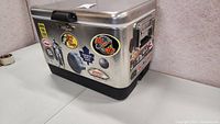 Front and right side view of metal Coleman cooler with decals and stickers on its sides.