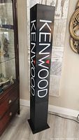 Full view of the tall black rectangular Kenwood advertising sign with illuminated logo, standing on floor near wall and furniture.