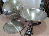 Electric wok with lid, perforated steamer basket, and temperature control dial plug shown from above on table.