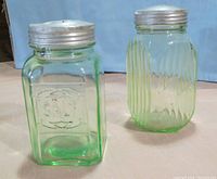 Two vintage green glass storage jars with metal screw tops, one square embossed and the other ribbed