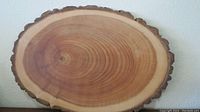 Oval wooden slab with natural bark edge and visible growth rings on wood surface against a light-colored wall.