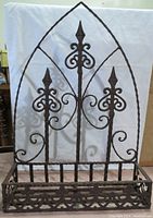 Photo of an ornate metal hanging shelf with a pointed arched top, decorated with fleur-de-lis styled vertical spikes and scrollwork, and a lattice pattern base for holding items.