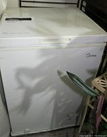 Front view of white Midea chest freezer with manual on top and plant leaves nearby.