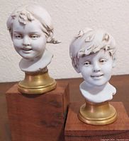 Front view of both miniature child busts with smooth white finish and golden bases on wooden blocks.