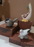 Three ceramic miniature bird figurines: gray sparrow, white goose with green leaf, and white dove with black accents, displayed on wooden blocks.