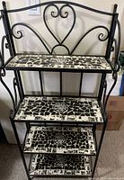 Front view of the black metal folding shelf with three mosaic tile shelves showing decorative scrollwork and heart-shaped back motif.