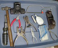 Photo showing variety of hand tools including hammer, screwdriver, pliers, saw, hand plane, chalk line reel, and drill bits case arranged on blue surface.