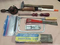 Visible tools include a hand-crank drill with wooden handle, a wooden handled hammer, a screwdriver with wooden handle, a sharpening stone in its box, a metal coping saw frame, and a wooden handled scraper or chisel tool.
