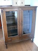Front view of antique wooden display cabinet showing two glass-paneled doors and two drawers below. Shows overall design and finish of the piece.