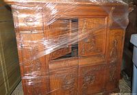 Front view of the antique sewing cabinet showing the intricate carved patterns on the doors and drawers wrapped in shrink wrap.