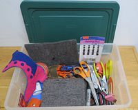 Photo showing plastic container with multiple pairs of scissors and other crafting tools, colorful felt pieces, and a package of sewing brads