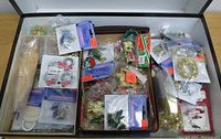Box filled with assorted small decorative miniature Christmas ornaments in original packaging, including bells, wreaths, and poinsettias.