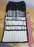 An assortment of varied sizes of knitting needles with plastic and metal construction organized in a black fabric holder and two red long needles beside it.