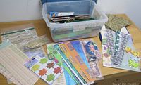 Wide view showing the plastic tub filled with an assortment of sticker sheets and papercraft items spread around it including various designs and colors