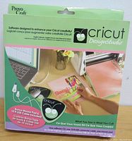 Front of Cricut DesignStudio software package showing branding, mouse pad, USB cable included, and a laptop with cutter interface.
