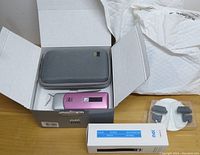 Open box containing a pink No No hair removal device, a grey carrying case, replacement blades in plastic packaging, a white instruction manual, and the original packaging box.