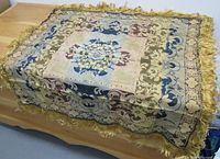 Image showing vintage boho style tablecloth with floral and scroll designs, edged with golden fringe, laid over a wooden table.