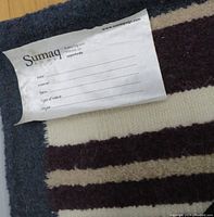 Close-up of Sumaq label on a striped wool rug showing brand and material details.