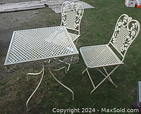 Full view of the white metal bistro table with two matching folding chairs on grass.