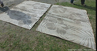 Two indoor/outdoor carpets laid out on grass showing floral and leaf patterns in neutral tones.