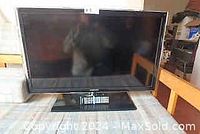 Black Samsung flat-screen television on a TV stand with remote control on base. Display visible, slight reflections.