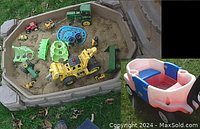 Sandbox containing sand and several toy construction vehicles including a yellow toy excavator, multiple green tractors, and other small toys. Adjacent is the Little Tykes 2-seat wagon in blue, red and white plastic.
