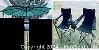 Photo showing open large outdoor umbrella with teal canopy and metal frame, alongside two black folding camp chairs on green grass.