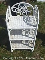 Front and top view of white painted wicker shelving unit with three shelves and decorative backrest design in outdoor grass setting.