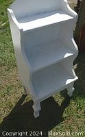 Image of 3-tier white painted wood shelf unit standing outdoors on grass, showing overall shape and carved legs.