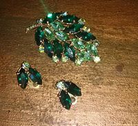 Rhinestone brooch with matching clip-on earrings shown on a wooden surface showing detailed view of green marquise and round rhinestones in gold-tone setting.