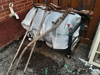 Wide shot showing collection of old garden tools outdoors including two hoes, a scythe wrapped with rope on handle, and shears all resting on top of a wrapped object.