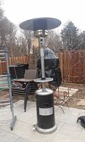 Full view of the freestanding gas patio heater showing the black and silver metal base, vertical column, and round top reflector on an outdoor patio.