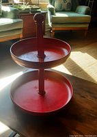 Front view of red two-tier metal farmhouse stand showing the top tier, central post, and bottom tier on the hardwood table.