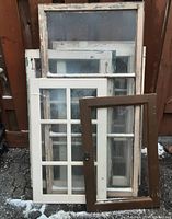 Seven old wooden window frames stacked against a fence, showing multiple finishes and glass pane styles.