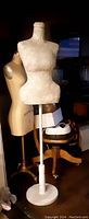 Photo showing full dress form on white wooden stand with round base, with two other dress forms and furniture in the background.