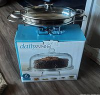 Silver metal chafing dish with glass lid resting on top of a blue and white Dailyware cake stand box containing the glass cake stand.