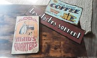 Three vintage decorative signs on a wooden surface, including one wooden Maid's Quarters sign and two metal signs.