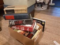 Two images of cardboard boxes filled with hardcover and paperback books of various titles and genres.