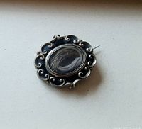 Front view showing ornate black enamel and gold accented frame with oval glass center containing woven hair.